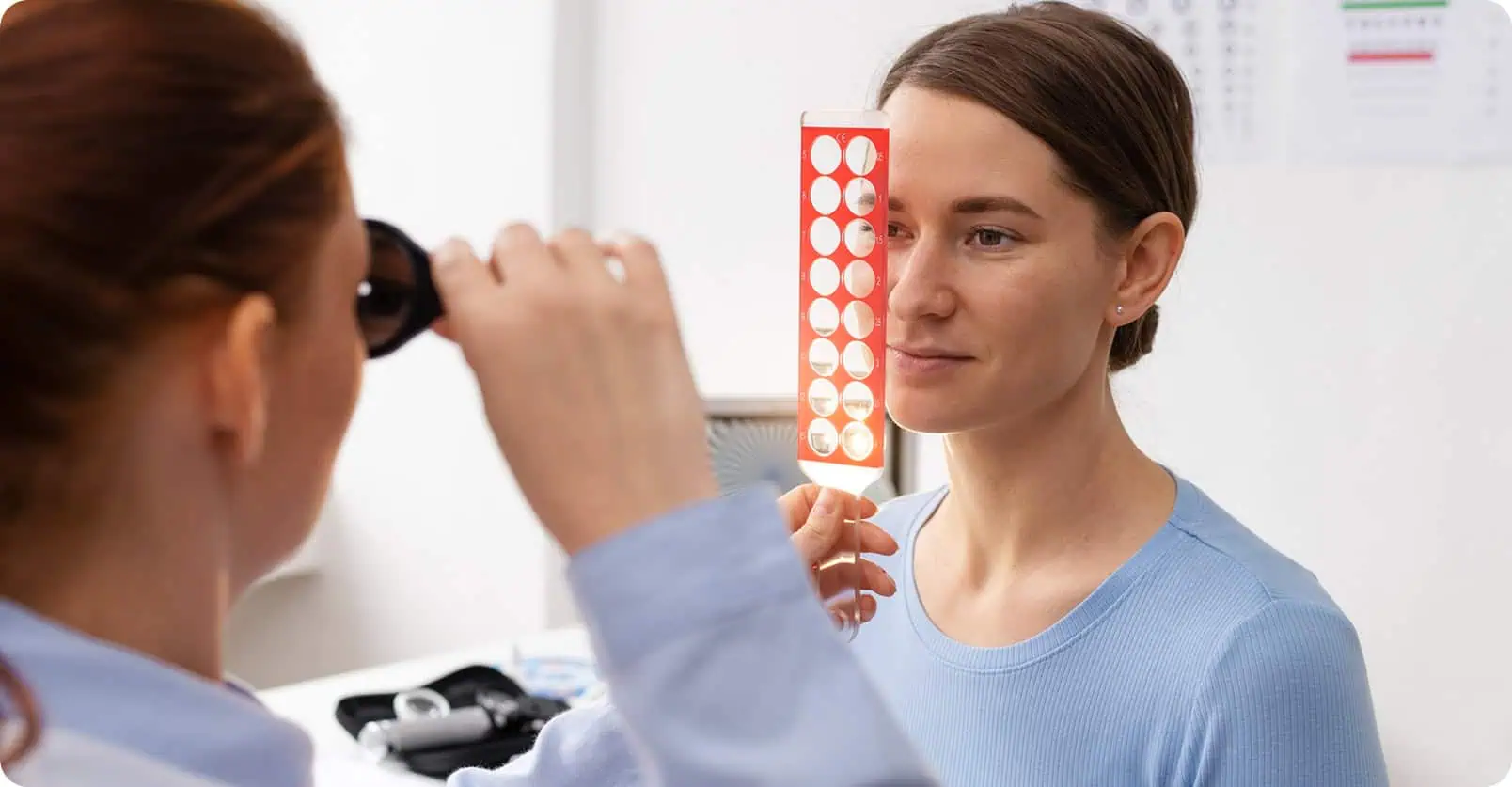 Optometrist examines woman's eye with eye chart in clinic setting.
