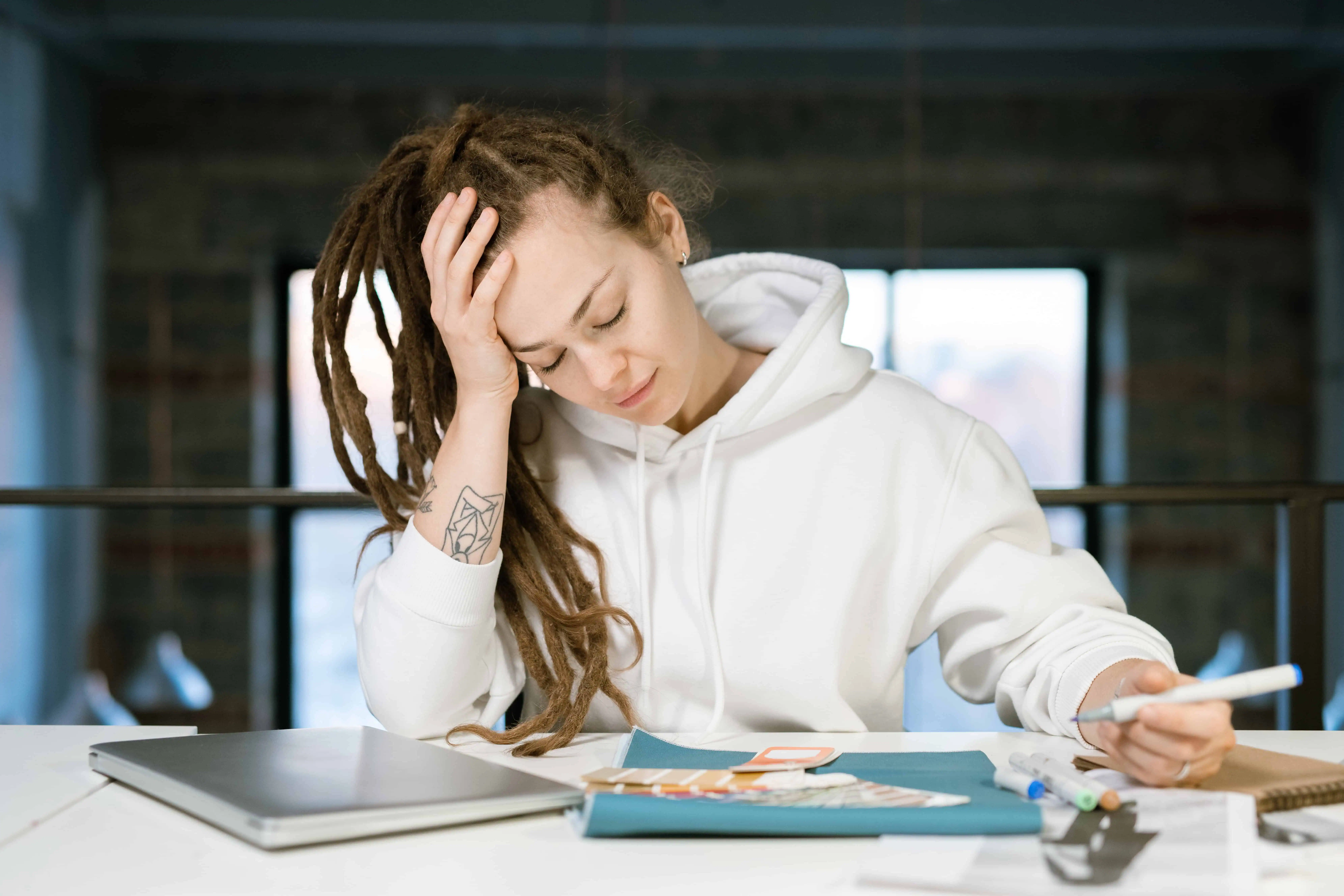 Young woman with dreadlocks looking stressed at desk with papers and laptop.