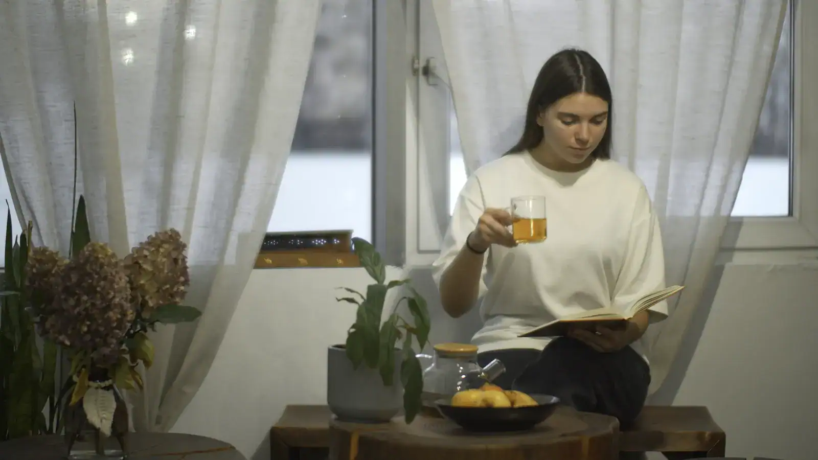 Woman drinking tea and reading a book by a window as part of an alcohol-free evening routine